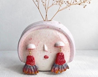 May include: A ceramic face-shaped vase in pale pink with speckled details, featuring a small nose and red lips. Two dangling earrings with textured red tops and multi-colored, fan-shaped bottoms hang from the vase. Dried white flowers are inside.