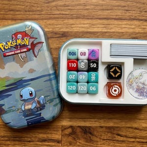 May include: A Pokémon Trading Card Game set, featuring a blue tin with Squirtle artwork. The set includes dice, a coin, and a compartment for cards. The dice are various colors and have numbers. The tin has the Pokémon Trading Card Game logo.