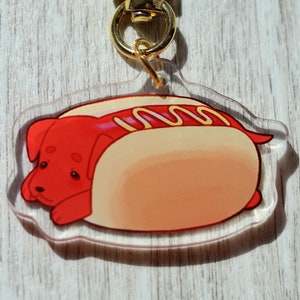 May include: A clear acrylic keychain featuring a cartoon dog shaped like a hot dog with mustard and ketchup. The keychain has a gold metal clasp.