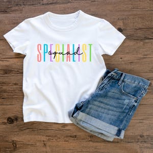 Puede incluir: Camiseta blanca con el texto "SPECIALIST squad" en letras coloridas. La palabra "squad" está escrita en cursiva. La camiseta se combina con pantalones cortos de mezclilla, todo sobre una superficie de madera.