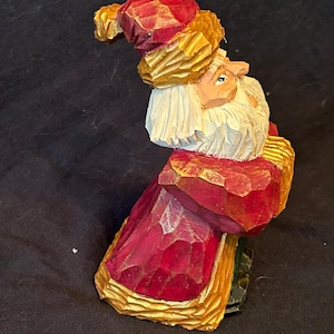 Puede incluir: Figura de madera tallada de Papá Noel. La figura viste una túnica roja con adornos dorados y un gorro a juego. Papá Noel tiene una larga barba blanca y una expresión sonriente. La talla tiene un aspecto texturizado y facetado.