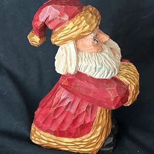 Puede incluir: Una figura de Santa Claus de madera tallada a mano con un traje rojo, ribetes dorados y una barba blanca. La figura está de pie con los brazos cruzados y mira hacia la derecha.