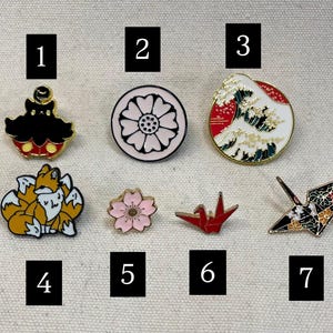 May include: Ten enamel pins with different designs. The pins feature a black and white creature, a red and white wave, a pink flower, a group of orange foxes, a pink flower, a red origami crane, a black and white origami crane, and numbers 1 through 7.