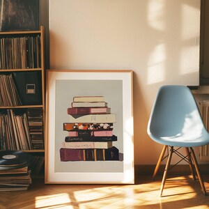 May include: A framed print of a stack of books with different coloured covers on a light grey background. The books are stacked in a pile, with the top book being a light beige colour. The other books are a mix of dark red, pink, and black.