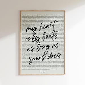 Puede incluir: Una impresión enmarcada con un marco de color madera clara. La impresión presenta un fondo de texto impreso con la cita manuscrita, "my heart only beats as long as yours does" en escritura negra. El autor y el título del libro están en la parte inferior.