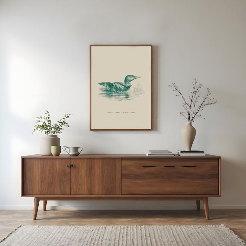 May include: A framed print featuring a teal illustration of a loon swimming on water. The artwork is set against a cream background and framed in a dark wood. The text "STUDIO CANADIAN WILD BIRD" is printed below the image. The print hangs above a mid-century modern wooden cabinet.