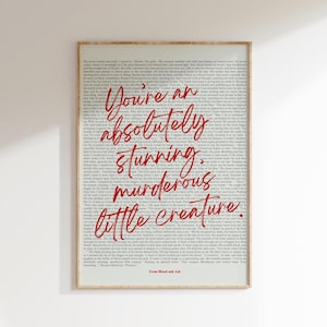 May include: A framed print with a cream-colored background filled with small text. Bold, red cursive text reads "You're an absolutely stunning, murderous little creature." The bottom of the print reads "From Blood and Ash."
