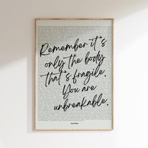 Puede incluir: Una impresión enmarcada con un marco de madera clara. La impresión presenta un fondo de texto impreso con las palabras "Remember it's only the body that's fragile. You are unbreakable." en un estilo manuscrito. El texto "Iron Flame" está en la parte inferior.