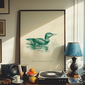 May include: A framed print featuring a teal illustration of a loon swimming in water. The print has the text "STUPID CANADIAN WOLF BIRD" below the image. The print is displayed on a table with a record player, lamp, and fruit.