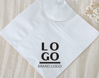 May include: White square paper napkin with the word "LOGO" in black, with "BRAND LOGO" underneath. A clear glass is on top of the napkin. The napkin is on a cream-colored surface.