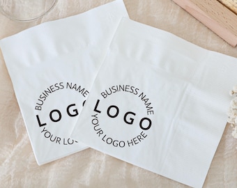 May include: Two white paper napkins with a circular logo design printed in black. The logo includes the words "BUSINESS NAME" and "LOGO" with the text "YOUR LOGO HERE" at the bottom. The napkins are on a light-colored surface.