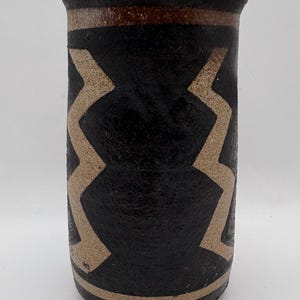 May include: A cylindrical ceramic vase with a black background and tan zigzag patterns. The top and bottom rims have a tan band, and the top rim also has a brown band. The vase has a textured surface.