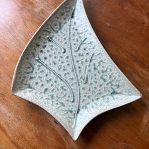 May include: A leaf-shaped ceramic dish featuring a textured, mottled glaze in shades of pale green and cream. The surface is decorated with a raised, branching vein pattern that extends from the base toward the pointed tip of the dish.