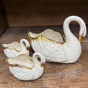 May include: A set of three white ceramic swan-shaped planters with gold trim. The swans have curved necks and open containers for plants or decor. The largest swan is in the back, with two smaller swans in front.