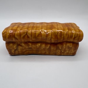 May include: A rectangular ceramic container with a brown, textured exterior. The container has a lid and is designed to resemble a loaf of bread. The ceramic piece has a glossy finish and is likely used for decorative or storage purposes.