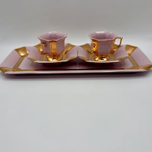 May include: A set of two pink teacups and saucers with gold trim, presented on a matching pink tray with gold accents. The teacups have a unique, angular design, and the set is arranged on a white background.