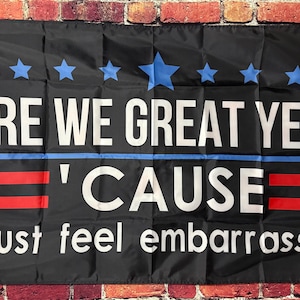 May include: Black flag with white text: "ARE WE GREAT YET?" and "'CAUSE I just feel embarrassed." The flag features blue stars, a blue stripe, and red stripes.