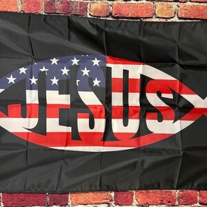 Op de afbeelding: Zwarte vlag met een vissymbool met het ontwerp van de Amerikaanse vlag en het woord "JESUS" in het wit. De vlag heeft een witte rand en wordt getoond tegen een bakstenen muur.