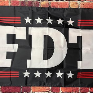 Peut inclure: Drapeau noir avec les lettres blanches "FDT" en gras. Le drapeau comporte cinq étoiles blanches au-dessus et en dessous des lettres, et des rayures rouges en haut et en bas.