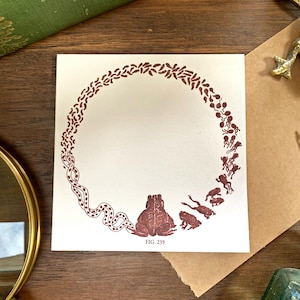 May include: An off-white square card featuring a brown illustration of a frog and a circular design of leaves and frog-like figures. The text "FIG. 239" is printed at the bottom. The card is on a wooden surface with a magnifying glass and other vintage items.
