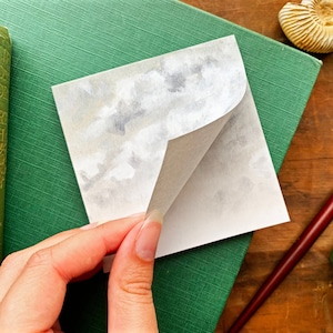 Puede incluir: Una nota adhesiva blanca con un diseño de acuarela gris en la parte delantera. La nota está parcialmente despegada para revelar la parte posterior blanca en blanco.