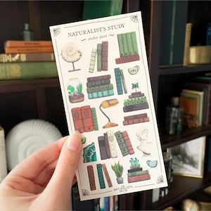 Op de afbeelding: Een vel stickers met illustraties van stapels boeken, een lamp, een fossiel en planten. Het vel stickers heeft de titel "Naturalist's Study - sticker sheet" en de tekst "Naturalist's Guild" onderaan.