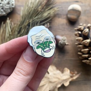 May include: A white enamel pin with a cartoon portrait of a man with a green butterfly on his nose. The butterfly has green wings and a black body.