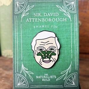 May include: A green enamel pin featuring a cartoon portrait of Sir David Attenborough with a green butterfly on his nose. The pin has white text that reads "Sir David Attenborough Enamel Pin" and "The Naturalist's Guild".