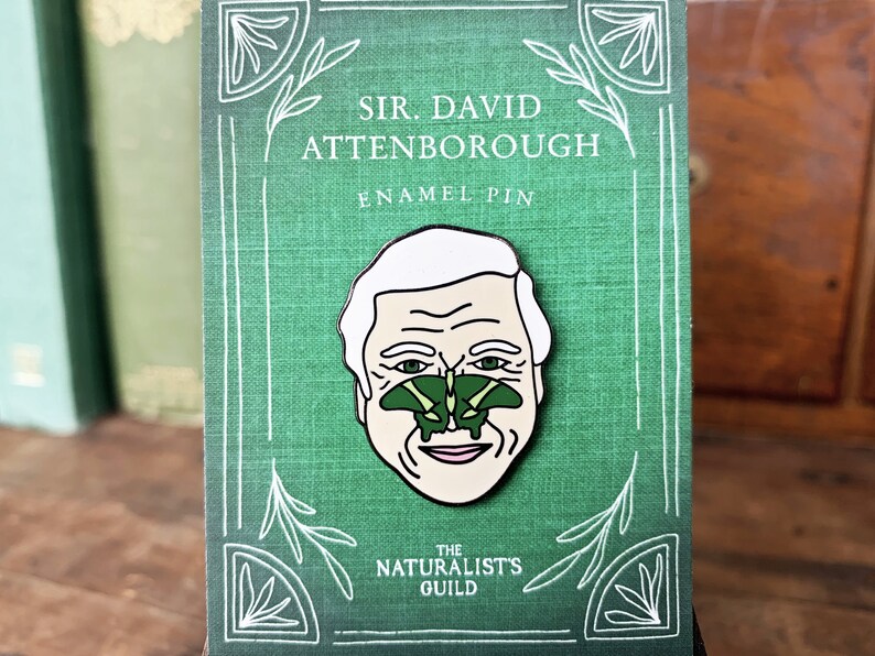 May include: A green enamel pin featuring a cartoon portrait of Sir David Attenborough with a green butterfly on his nose. The pin has white text that reads "Sir David Attenborough Enamel Pin" and "The Naturalist's Guild".