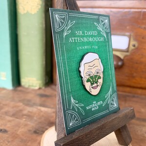 May include: A green enamel pin featuring a portrait of Sir David Attenborough with a green butterfly on his nose. The pin is displayed on a small wooden easel. The text "SIR. DAVID ATTENBOROUGH ENAMEL PIN THE NATURALISTS GUILD" is printed on the card behind the pin.