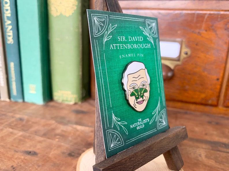 May include: A green enamel pin featuring a portrait of Sir David Attenborough with a green butterfly on his nose. The pin is displayed on a small wooden easel. The text "SIR. DAVID ATTENBOROUGH ENAMEL PIN THE NATURALISTS GUILD" is printed on the card behind the pin.