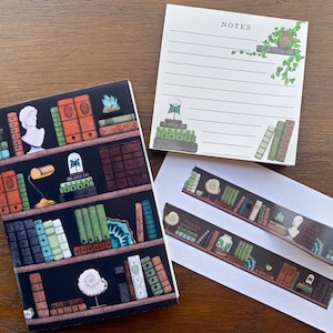 May include: A set of three stationery items featuring a bookshelf design. The set includes a small notebook, a notepad with the word "NOTES" at the top, and a sheet of decorative washi tape. The bookshelf design features a variety of books, plants, and decorative objects.