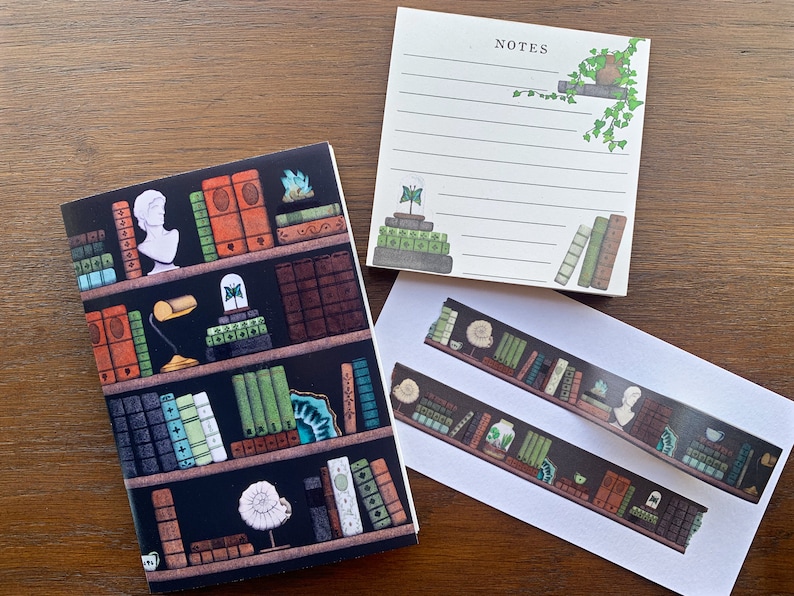 May include: A set of three stationery items featuring a bookshelf design. The set includes a small notebook, a notepad with the word "NOTES" at the top, and a sheet of decorative washi tape. The bookshelf design features a variety of books, plants, and decorative objects.