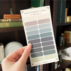 May include: A sticker sheet with 15 labels in various shades of blue and grey. The sheet is titled "Dark Academia Labels" and has the text "sticker sheet" below the title. The sheet is on a wooden shelf with books behind it.