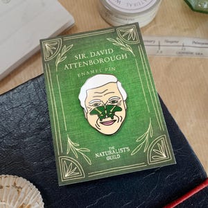 May include: A green enamel pin featuring a portrait of Sir David Attenborough with a green butterfly on his nose. The pin has the text "Sir David Attenborough Enamel Pin" and "The Naturalists Guild" on it.