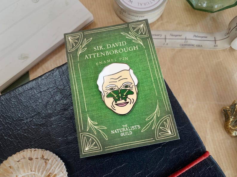 May include: A green enamel pin featuring a portrait of Sir David Attenborough with a green butterfly on his nose. The pin has the text "Sir David Attenborough Enamel Pin" and "The Naturalists Guild" on it.