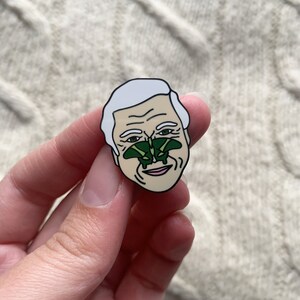 May include: A white enamel pin featuring a cartoon portrait of a man with a green butterfly on his nose.