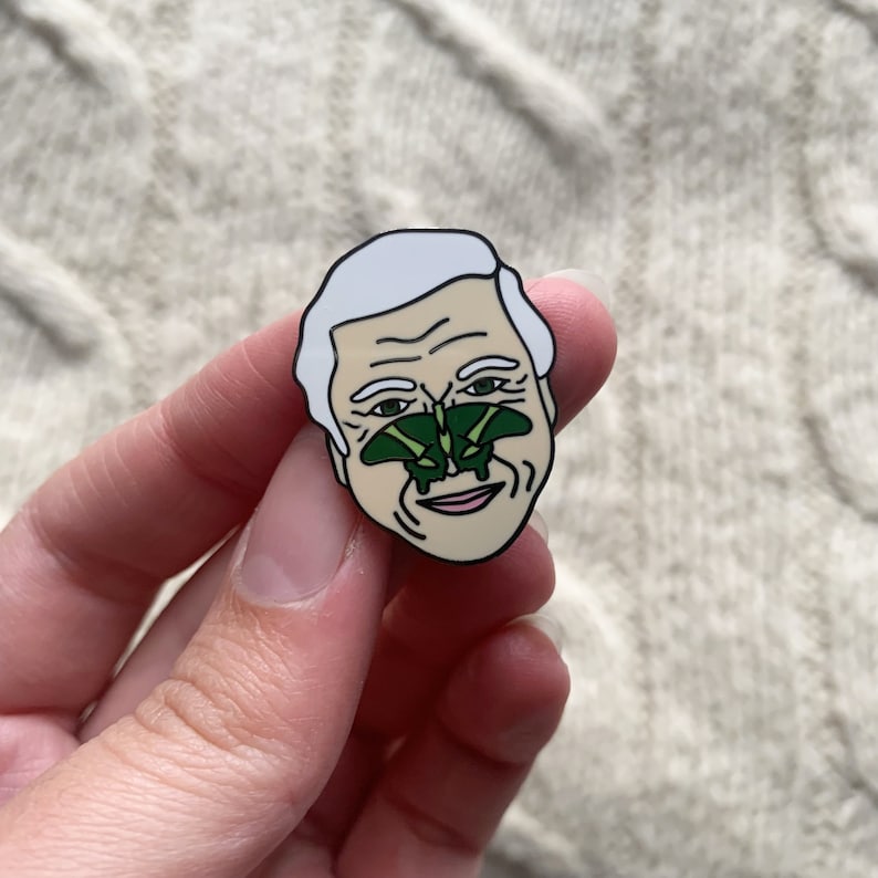 May include: A white enamel pin featuring a cartoon portrait of a man with a green butterfly on his nose.