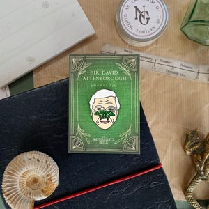 May include: A green enamel pin featuring a portrait of Sir David Attenborough with a green butterfly on his nose. The pin is surrounded by a green border with the text "Sir. David Attenborough Enamel Pin" and "The Naturalists Guild".