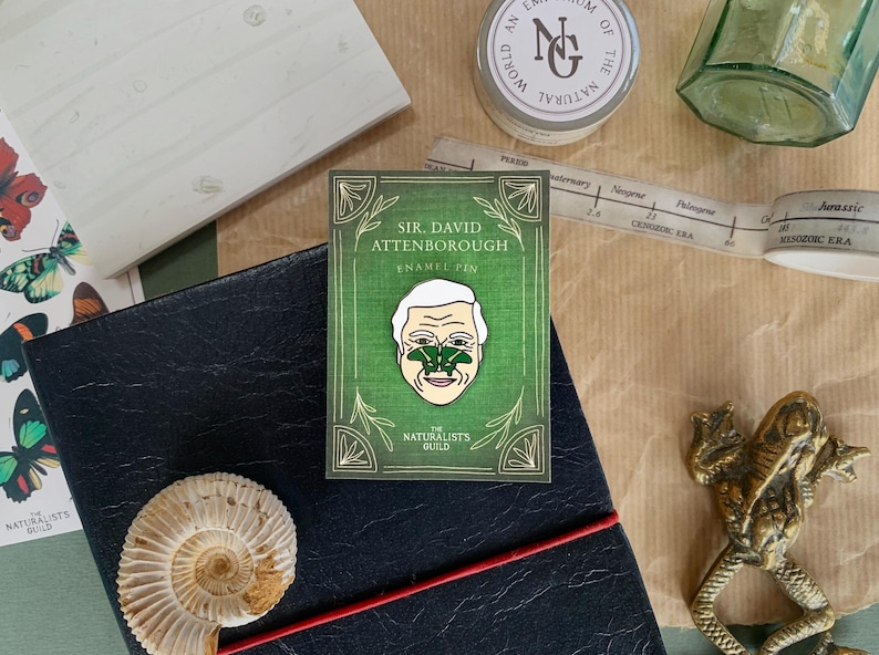 May include: A green enamel pin featuring a portrait of Sir David Attenborough with a green butterfly on his nose. The pin is surrounded by a green border with the text "Sir. David Attenborough Enamel Pin" and "The Naturalists Guild".
