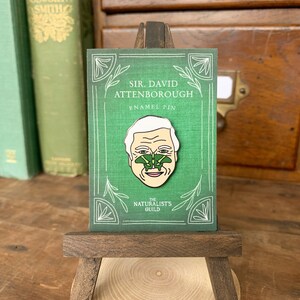 May include: A green enamel pin featuring a portrait of Sir David Attenborough with a green butterfly on his nose. The pin is on a small wooden easel and has the text "Sir David Attenborough Enamel Pin" and "The Naturalists Guild" on it.