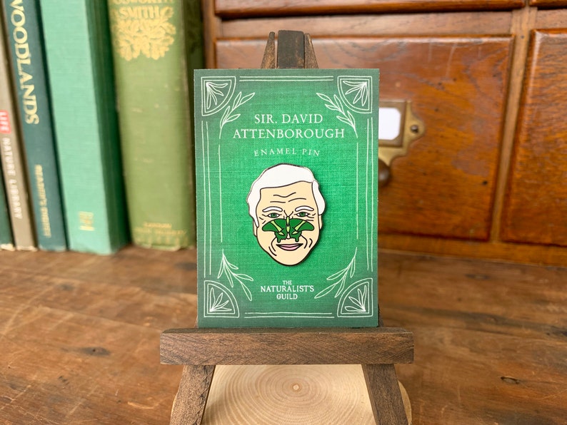 May include: A green enamel pin featuring a portrait of Sir David Attenborough with a green butterfly on his nose. The pin is on a small wooden easel and has the text "Sir David Attenborough Enamel Pin" and "The Naturalists Guild" on it.