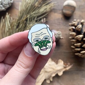 May include: A white enamel pin of a man's face with two green butterflies on his nose.