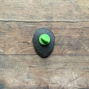 May include: The back of a black enamel pin with a green plastic pin back.