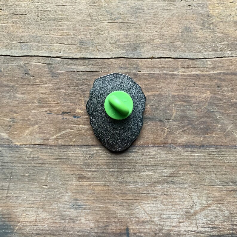 May include: The back of a black enamel pin with a green plastic pin back.