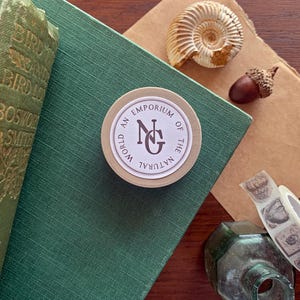 May include: A still life arrangement includes a green book, a round sticker with the text "World An Emporium of the Natural", a seashell, an acorn, and decorative tape. The items are arranged on a green surface and a brown paper.