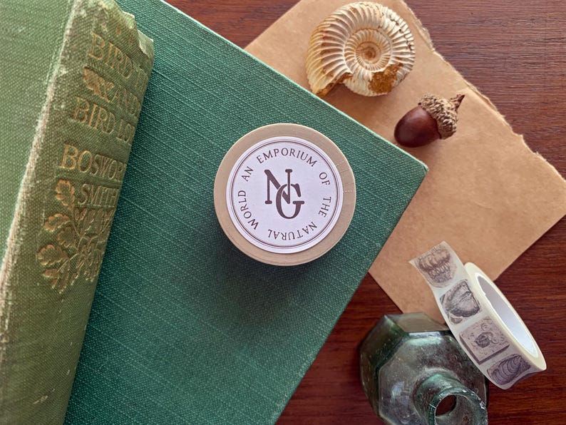 May include: A still life arrangement includes a green book, a round sticker with the text "World An Emporium of the Natural", a seashell, an acorn, and decorative tape. The items are arranged on a green surface and a brown paper.