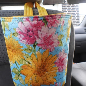 May include: A blue and yellow floral fabric tote bag with yellow handles. The bag is hanging from the headrest of a car seat.