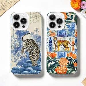 May include: Two phone cases with distinctive designs. One displays a tiger in a blue and white floral pattern, the other features a tiger against a backdrop of orange flowers and blue tiles. Both cases have a clear border and a camera cutout.