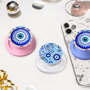 Op de afbeelding: Verschillende telefoonhouders met een evil eye-motief. De houders zijn in verschillende kleuren, waaronder blauw, wit en roze. Eén is bevestigd aan een telefoon met een bloemenhoesje. De achtergrond bevat een discobal.